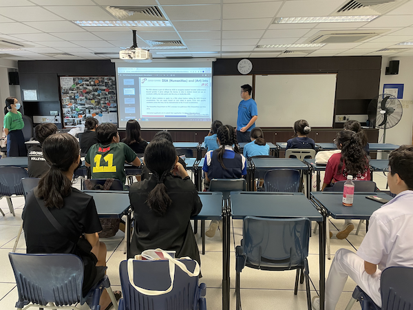 A student in a blue shirt presents a slideshow to a classroom of seated students facing a projector screen.